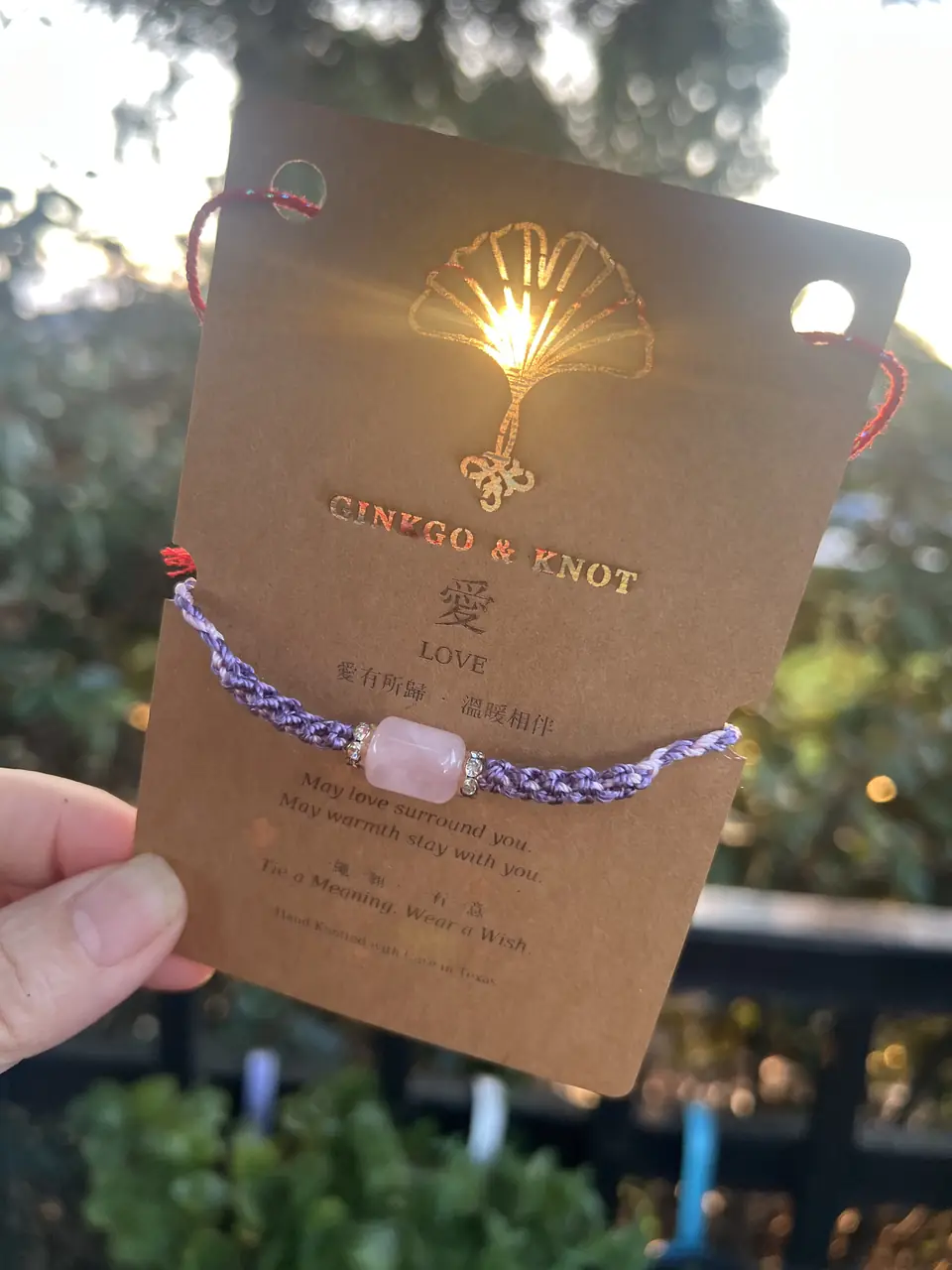 A Ginkgo & Knot blessing card held up to the setting sun, light shining through the cut-out ginkgo above a rose quartz bracelet.